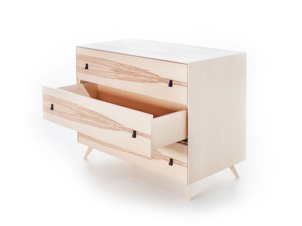 Moderne Kommode mit heller Holzoberfläche und dezenter Maserung. Drei großzügige Laden mit je zwei kleinen Lederlaschen als Griffen. Die mittlere Lade ist halb geöffnet. Die Kommode steht auf vier grazilen, leicht schrägen Füßen. Das Design wirkt minimalistisch, klar und hochwertig.