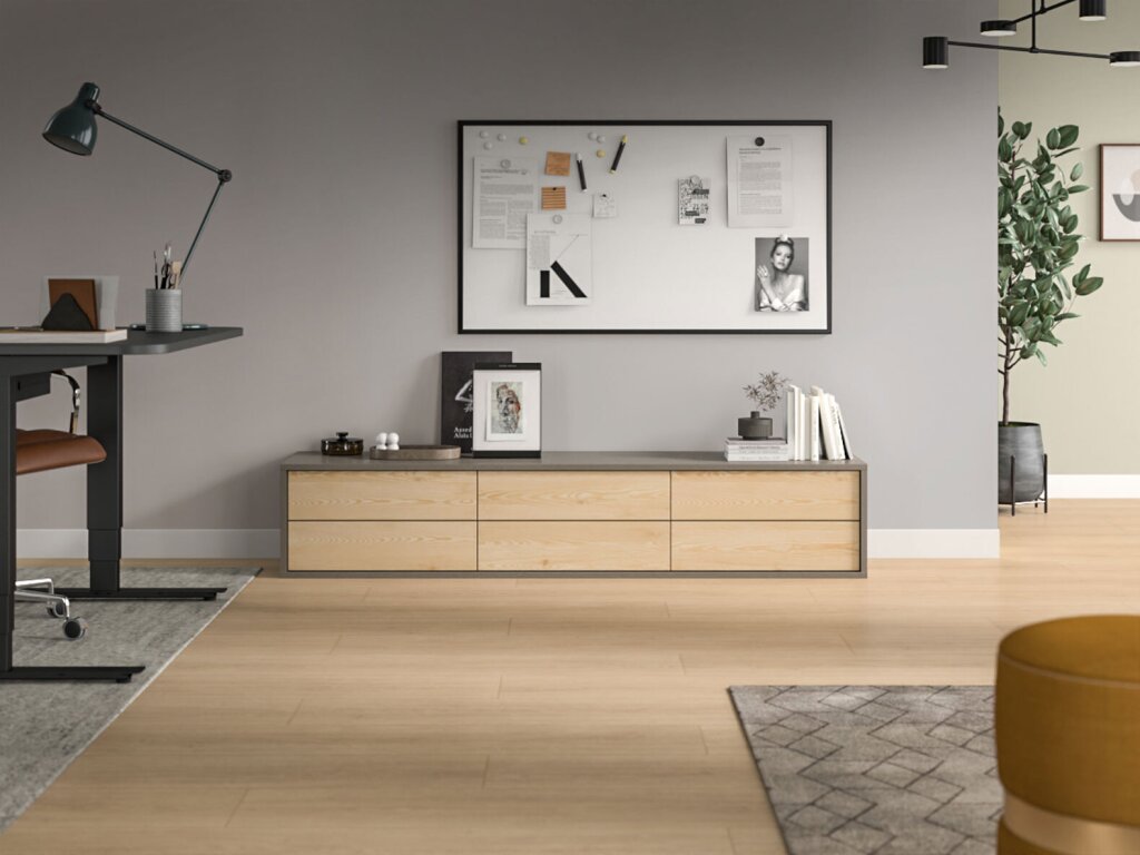 Lowboard mit sechs grifflosen Laden in heller Holzoptik und klarer, geradliniger Formensprache. Das Möbelstück steht vor einer grau gestrichenen Wand und wird von dekorativen Objekten wie Büchern, einem gerahmten Bild und einer kleinen Vase ergänzt. Darüber hängt ein großes, gerahmtes Whiteboard mit Notizen und Fotos. Links im Bild befindet sich ein höhenverstellbarer Schreibtisch mit schwarzem Gestell, rechts sorgt eine Pflanze in schwarzem Topf für einen natürlichen Akzent. Der Raum wirkt hell, modern und aufgeräumt, mit warmem Holzboden und sanften Farben.