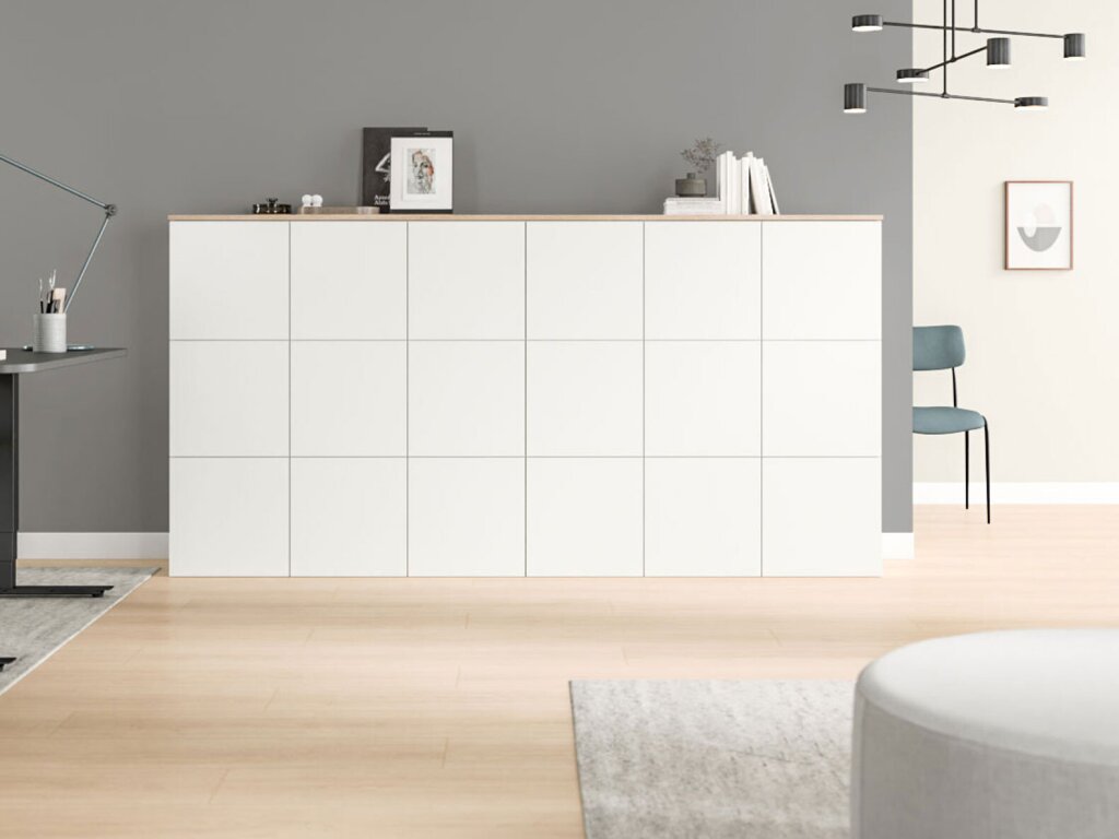 An einer grauen Wand eines Büros steht ein großes Highboard mit 18 fast quadratischen Elementen - jeweils sechs in drei Reihen angeordnet. Das Möbel hat weiße Fronten und eine Abdeckplatte aus hellem Holzdekor. Auf dem Highboard stehen Bücher, ein Bild und kleinere Dekorationsartikel.