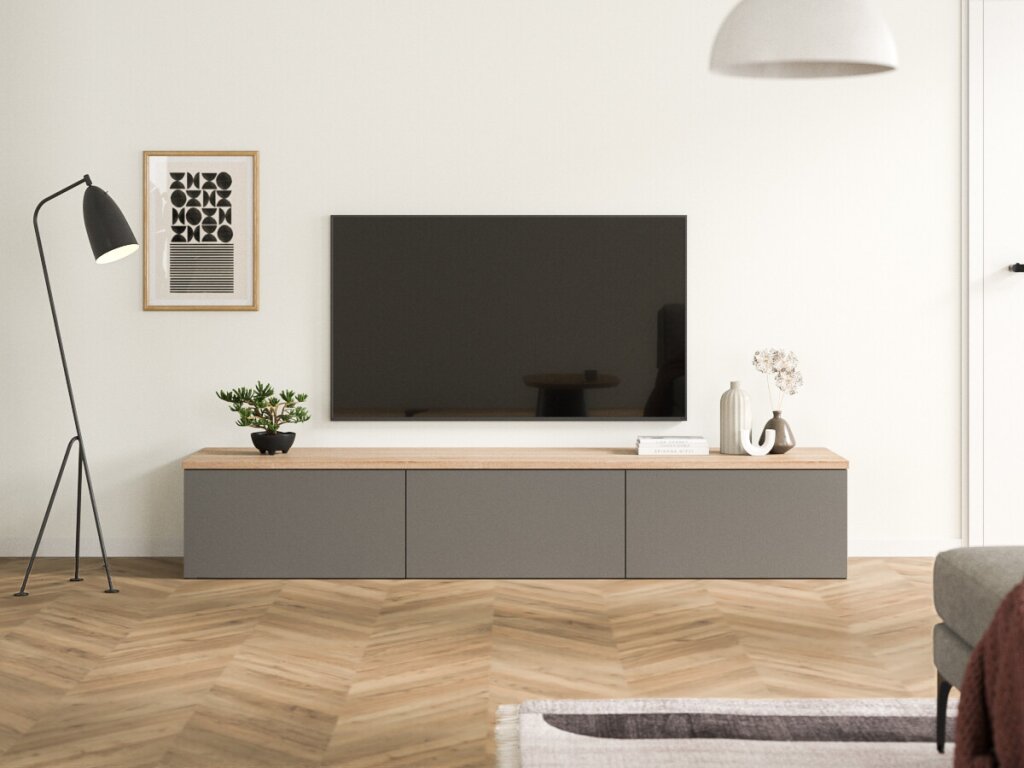Ein stehendes Sideboard aus grauen Dekorplatten mit einer Deckplatte aus Eichenholz. Das TV-Board hat drei Segmente, die als Schubladen, Drehtüren oder Klapptüren gearbeitet werden können. Das Möbel lässt sich im Möbelplaner entsprechend anpassen. Das Board steht in einem schlichten Wohnzimmer, links eine moderne Lampe mit zartem Stahlgestell, rechts ein angeschnittener Wohnzimmersessel und eine weiße Tür. Dezente Dekoration und ein abstraktes Wandbild runden das Bild ab.